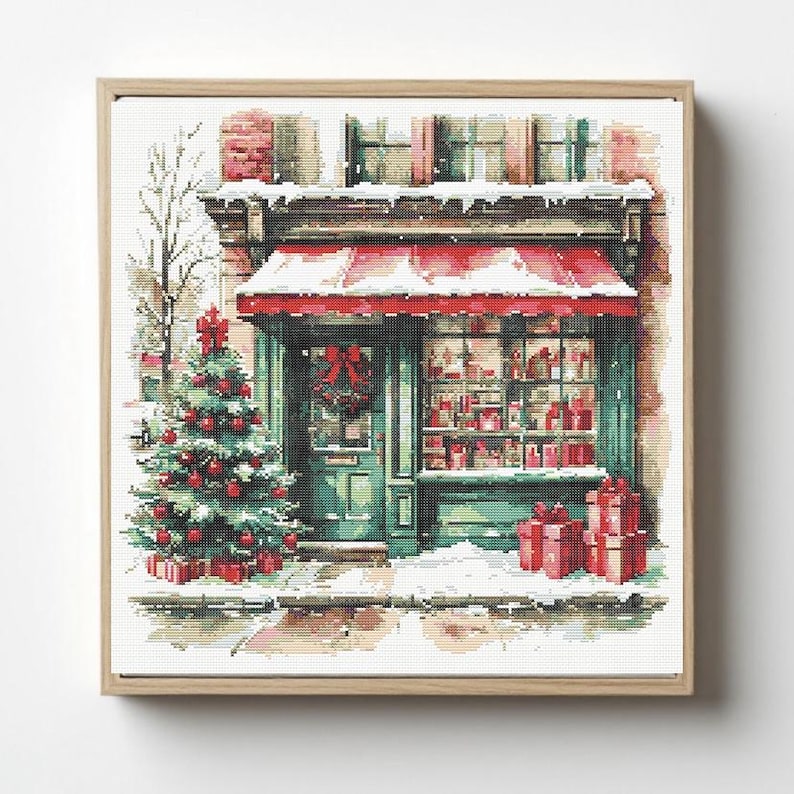 May include: A cross-stitch pattern depicting a storefront with a red awning, a decorated Christmas tree with red ornaments, and wrapped gifts. The scene features snow on the ground and building ledges, with a wreath on the door and a window display.