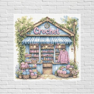 Könnte beinhalten: Aquarellillustration eines Häkelladens mit blauem Vordach und dem Wort "Crochet" über dem Eingang. Regale sind mit Garn und fertigen Artikeln gefüllt. Körbe mit Garn und Blumen befinden sich vor dem Geschäft. Ein rosa Pullover hängt zur Ansicht.