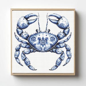 May include: A square cross-stitch embroidery of a crab centered on a white background, framed in light wood. The crab features intricate blue and white floral patterns across its shell and claws, mimicking traditional porcelain ceramic designs.