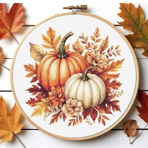 Può includere: Un telaio da ricamo circolare in legno contenente un disegno a punto croce di due zucche, una arancione e una bianca, circondate da un grappolo di foglie e fiori dai colori autunnali nelle tonalità del rosso, arancione, giallo e marrone.