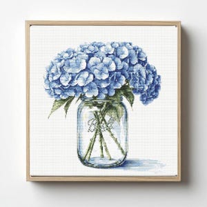 Puede incluir: Impresión enmarcada de un ramo de hortensias azules en un tarro de cristal transparente. El tarro tiene la palabra "Ball" impresa. La obra de arte está sobre un fondo blanco y enmarcada en madera clara.