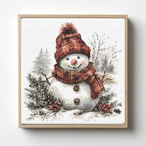 May include: A cross-stitch embroidery of a snowman wearing a red and brown plaid knit hat with a pom-pom and a matching plaid scarf. The snowman has a carrot nose, two brown buttons on its torso, and is surrounded by pine branches and red berries.