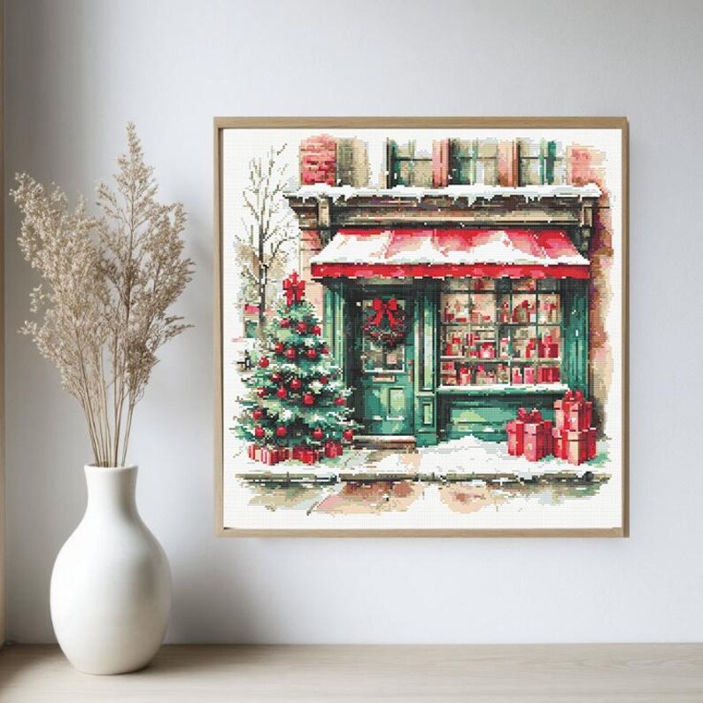 May include: A square cross-stitch pattern depicting a storefront with a red awning, a green door with a wreath, and a decorated Christmas tree outside. The scene features snow-covered ground, red gift boxes, and a brick building facade.