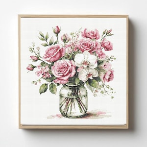 May include: Cross-stitch picture of a floral arrangement in a glass jar. The bouquet includes pink roses, white orchids, and foliage. The artwork is framed in a light brown wooden frame, set against a white background.