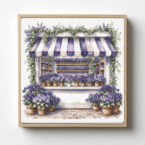 May include: A framed cross-stitch depicting a flower shop with a white and purple striped awning. The shop is filled with potted purple flowers and greenery. The image is set against a white background.