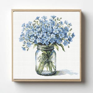 May include: A framed cross-stitch artwork featuring a bouquet of light blue forget-me-nots in a clear glass jar. The jar is tied with a twine bow and sits on a white background. The frame is made of light-colored wood.