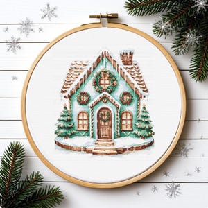 Puede incluir: Un bordado de punto de cruz de una escena invernal con una casa de jengibre turquesa con tejado nevado y coronas. La casa tiene una puerta y ventanas marrones, con dos pequeños árboles de Navidad a cada lado. El bordado está en un bastidor de madera.