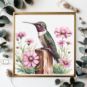 May include: A framed cross-stitch embroidery featuring a hummingbird with iridescent green and purple feathers perched on a wooden post, surrounded by pink cosmos flowers with green stems and leaves against a plain white background.