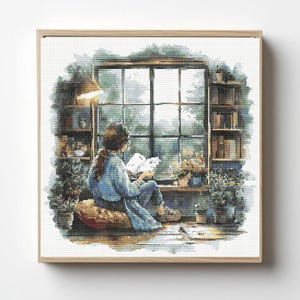 May include: A cross-stitch pattern featuring a person sitting on a cushion by a window, reading a book. The scene includes bookshelves, potted plants, and a view of trees outside, all framed in a light-coloured wooden square frame.