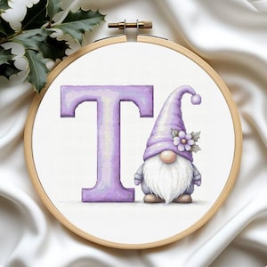 May include: Cross-stitch embroidery in a wooden hoop features a lavender letter "T" and a gnome. The gnome wears a lavender hat with a flower and has a long white beard. The background is white fabric.