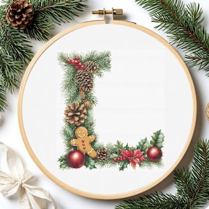 May include: Cross-stitch embroidery of the letter "L" decorated with Christmas elements. The design includes pine needles, pine cones, red berries, a gingerbread man, red ornaments, and poinsettias, all set within a wooden embroidery hoop.