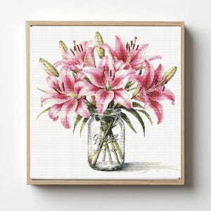 Op de afbeelding: Kruissteekkunst van roze lelies in een heldere glazen pot. De bloemen hebben witte accenten en groene bladeren. De pot staat tegen een witte achtergrond en is omlijst in licht hout.