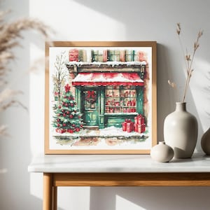 May include: A square framed print depicting a storefront with a red awning, a decorated Christmas tree, and snow-covered ground. The shop features green trim, a wreath on the door, and a window display filled with red gift boxes and presents.