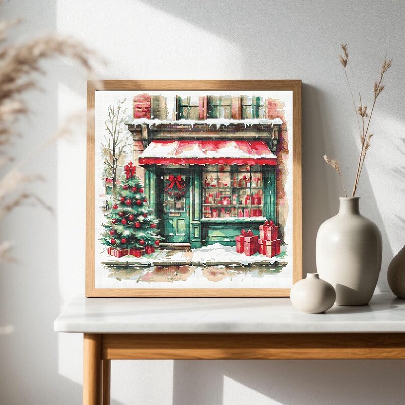 May include: A square framed print depicting a storefront with a red awning, a decorated Christmas tree, and snow-covered ground. The shop features green trim, a wreath on the door, and a window display filled with red gift boxes and presents.