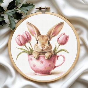 Könnte beinhalten: Kreuzstich-Stickerei eines Hasen in einer rosa Teetasse mit rosa Tulpen. Der Hase hat lange Ohren und ist hellbraun. Die Tasse hat ein Blumenmuster und ist in einem hölzernen Stickrahmen angeordnet.