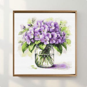 Op de afbeelding: Een kruissteekkunstwerk van een boeket paarse bloemen in een heldere glazen pot. De bloemen hebben groene bladeren en staan tegen een witte achtergrond. Het kunstwerk is ingelijst in een houten lijst.