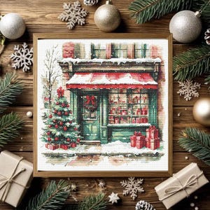 May include: A framed cross-stitch pattern depicting a storefront with a red awning, a decorated Christmas tree, and wrapped gifts. The scene features a green door with a wreath, snow-covered ledges, and a window display filled with red items.