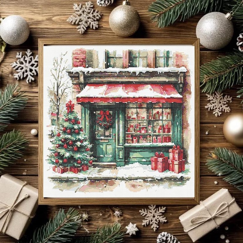 May include: A framed cross-stitch pattern depicting a storefront with a red awning, a decorated Christmas tree, and wrapped gifts. The scene features a green door with a wreath, snow-covered ledges, and a window display filled with red items.