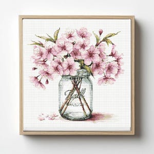Op de afbeelding: Kruissteekkunst van roze kersenbloesems in een heldere Mason jar, tegen een witte achtergrond. Op de pot staat het woord "MASON". Het kunstwerk is ingelijst in een lichtbruine houten lijst, wat een delicate en bloemige esthetiek creëert.