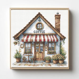 May include: A cross-stitch pattern depicting a coffee shop with a red and white striped awning, a brick chimney, and a round window. The building features a wooden door, two front windows, and potted plants on a stone patio in a light wood frame.
