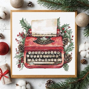 Può includere: Ricamo a punto croce di una macchina da scrivere rossa vintage con un foglio di carta bianco, circondata da agrifoglio, bacche e pigne. L'opera è incorniciata in una cornice di legno, con decorazioni natalizie e verde sullo sfondo.