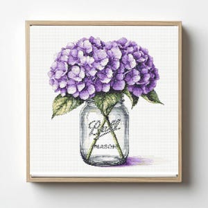 May include: Cross-stitch art of purple hydrangea flowers in a Ball Mason jar. The jar is clear with the Ball Mason logo. The artwork is framed in a light wood frame, set against a white background.