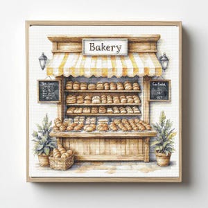 May include: A framed watercolor print of a bakery storefront. The bakery has a striped yellow and white awning, a sign that says "Bakery", and shelves filled with various baked goods. Two potted plants flank the bakery. Two black chalkboards are on either side of the bakery.
