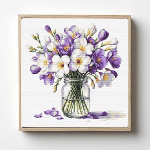 Op de afbeelding: Kruissteekkunstwerk van een boeket paarse en witte bloemen in een heldere glazen pot. De bloemen hebben gele centra en zijn gerangschikt met groene stelen. Het kunstwerk is ingelijst in een lichtbruine houten lijst.
