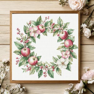 May include: Cross-stitch artwork of a wreath featuring red apples, pink and white blossoms, green leaves, and red berries. The wreath is set against a white background and framed in a wooden frame. The artwork is a detailed, close-up view.