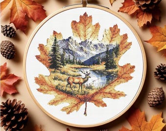 May include: A cross-stitch embroidery in a wooden hoop, featuring a detailed autumn landscape within a maple leaf design. The scene includes a deer, mountains, and a river, with fall foliage and pine cones surrounding the hoop.