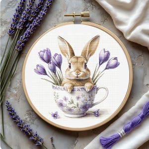 May include: A cross-stitch embroidery hoop featuring a brown bunny in a teacup with purple flowers. The teacup has a floral design. Lavender sprigs and purple embroidery floss are arranged around the hoop.