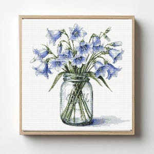 May include: A framed cross-stitch artwork featuring a bouquet of bluebell flowers in a clear glass mason jar. The flowers have delicate blue petals and green stems. The jar sits on a white background, and the frame is made of light-colored wood.