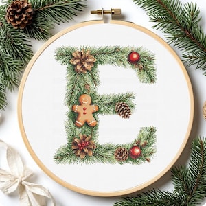 Puede incluir: Un bastidor de bordado con punto de cruz con la letra "E" adornada con decoraciones navideñas. Incluye follaje verde, piñas, un hombre de jengibre y bolas rojas. El bastidor de madera está sobre un fondo blanco.