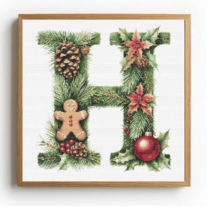 May include: Cross-stitch artwork of the letter "H" decorated with Christmas elements. The letter is formed from green pine needles and adorned with a pinecone, gingerbread man, red berries, poinsettias, and a red ornament. The artwork is framed in a wooden frame.