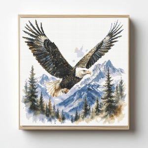 May include: A square cross-stitch embroidery piece featuring a bald eagle with wings spread wide in flight. The eagle has dark brown feathers and a white head and tail. Below the bird are blue-toned mountain peaks and green pine trees.