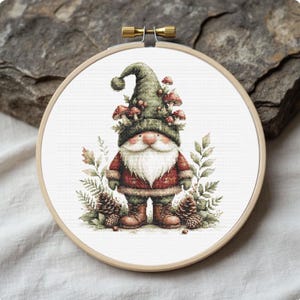 Puede incluir: Un bordado de punto de cruz en un aro de madera presenta un gnomo. El gnomo lleva un gorro verde adornado con setas, un abrigo rojo y botas marrones. Piñas y follaje rodean al gnomo, creando una escena encantadora y rústica.