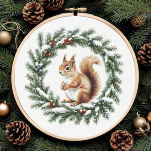 May include: A circular wooden embroidery hoop containing a cross-stitch pattern of a brown squirrel centered within a wreath of green pine branches, red berries, and white snow. The design is stitched on white fabric with a detailed finish.