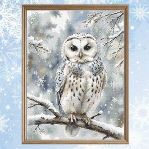 May include: A framed cross-stitch of a snowy owl perched on a snow-covered branch. The owl has white and gray feathers and is set against a winter forest scene with falling snowflakes. The frame is a light brown color.