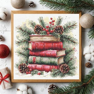May include: A square framed cross-stitch embroidery depicting a stack of four books in red and green hues, surrounded by pine branches, pinecones, and red berries. The artwork is set against a white background within a light wood frame.