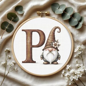 May include: Cross-stitch embroidery in a wooden hoop. The design features the letter "P" in brown and a gnome with a long white beard and a brown hat adorned with flowers. Eucalyptus and white flowers surround the hoop.