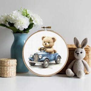 May include: A circular wooden embroidery hoop containing a cross-stitch pattern of a light brown teddy bear sitting in a vintage-style blue car. The bear has dark eyes and a small nose, while the car features round headlights and black wheels.