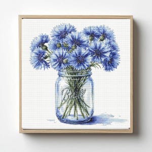 May include: A framed print featuring a bouquet of blue cornflowers in a clear glass mason jar. The jar has the word "Ball" printed on it. The flowers are a vibrant blue, and the background is white.
