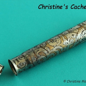 May include: A silver and gold metal tube with an intricate engraved design. The tube is open at both ends and has a cap on one end. The tube is on a turquoise background.