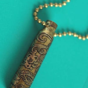 May include: A gold-colored bullet casing necklace with an intricate floral design. The necklace is made of a chain with small gold beads.