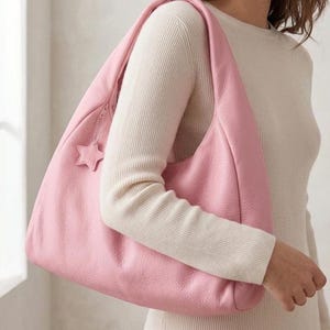 K&ouml;nnte beinhalten: Eine weiche, rosafarbene Hobo-Schultertasche mit strukturierter Oberfl&auml;che und einem breiten Einzelriemen. Die Tasche verf&uuml;gt &uuml;ber einen Rei&szlig;verschluss an der Oberkante und einen kleinen, passenden sternf&ouml;rmigen Anh&auml;nger an der Rei&szlig;verschlusslasche.