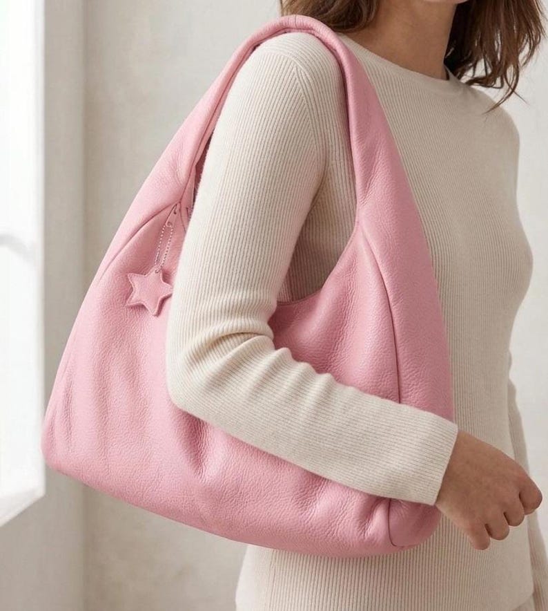 K&ouml;nnte beinhalten: Eine weiche, rosafarbene Hobo-Schultertasche mit strukturierter Oberfl&auml;che und einem breiten Einzelriemen. Die Tasche verf&uuml;gt &uuml;ber einen Rei&szlig;verschluss an der Oberkante und einen kleinen, passenden sternf&ouml;rmigen Anh&auml;nger an der Rei&szlig;verschlusslasche.