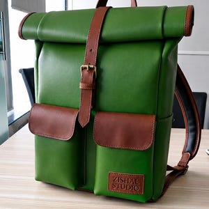 Puede incluir: Una mochila verde de cierre enrollable con detalles en cuero marrón, una correa vertical central con hebilla de latón, dos bolsillos delanteros con solapa y un parche de cuero rectangular con el texto ZISHAL STUDIO en el bolsillo derecho.