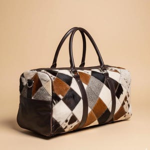 May include: A rectangular duffel bag with a patchwork design. The bag features a mix of brown, black, and white cowhide patches in a diamond pattern. It has dark brown leather handles and accents, with a matching leather base.