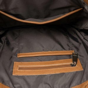 以下が含まれることがあります： ダークブラウンの布製ライニングが施されたタンカラーのハンドバッグの内側。バッグの上部にはオープンポケットがあり、その下にはタンカラーのレザーアクセントと金属製のジッパープルが付いたファスナー付きポケットが備わっています。