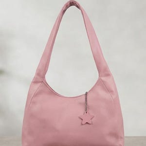 K&ouml;nnte beinhalten: Eine altrosa Hobo-Tasche mit glatter, matter Textur und einem einzelnen Schulterriemen. Die Tasche ist mit einem kleinen, f&uuml;nfzackigen Sternanh&auml;nger versehen, der an einer kurzen silberfarbenen Metallkette am oberen Rand befestigt ist.